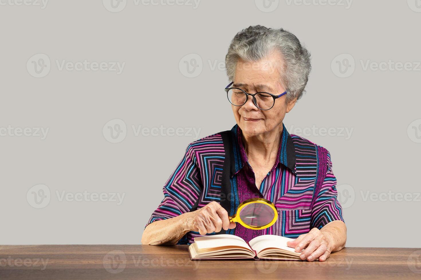 Senior woman is holding a magnifying glass for looking at a book while sitting at the table photo