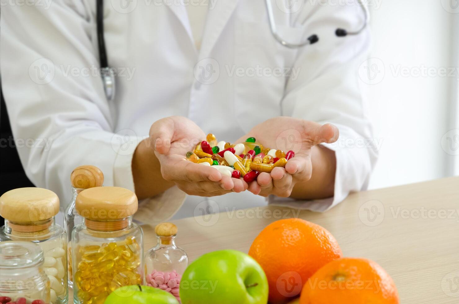Nutritionist doctor in white coat with stethoscope holding colorful pills in open palms. Jars of pills, apple and orange on clinic desk photo