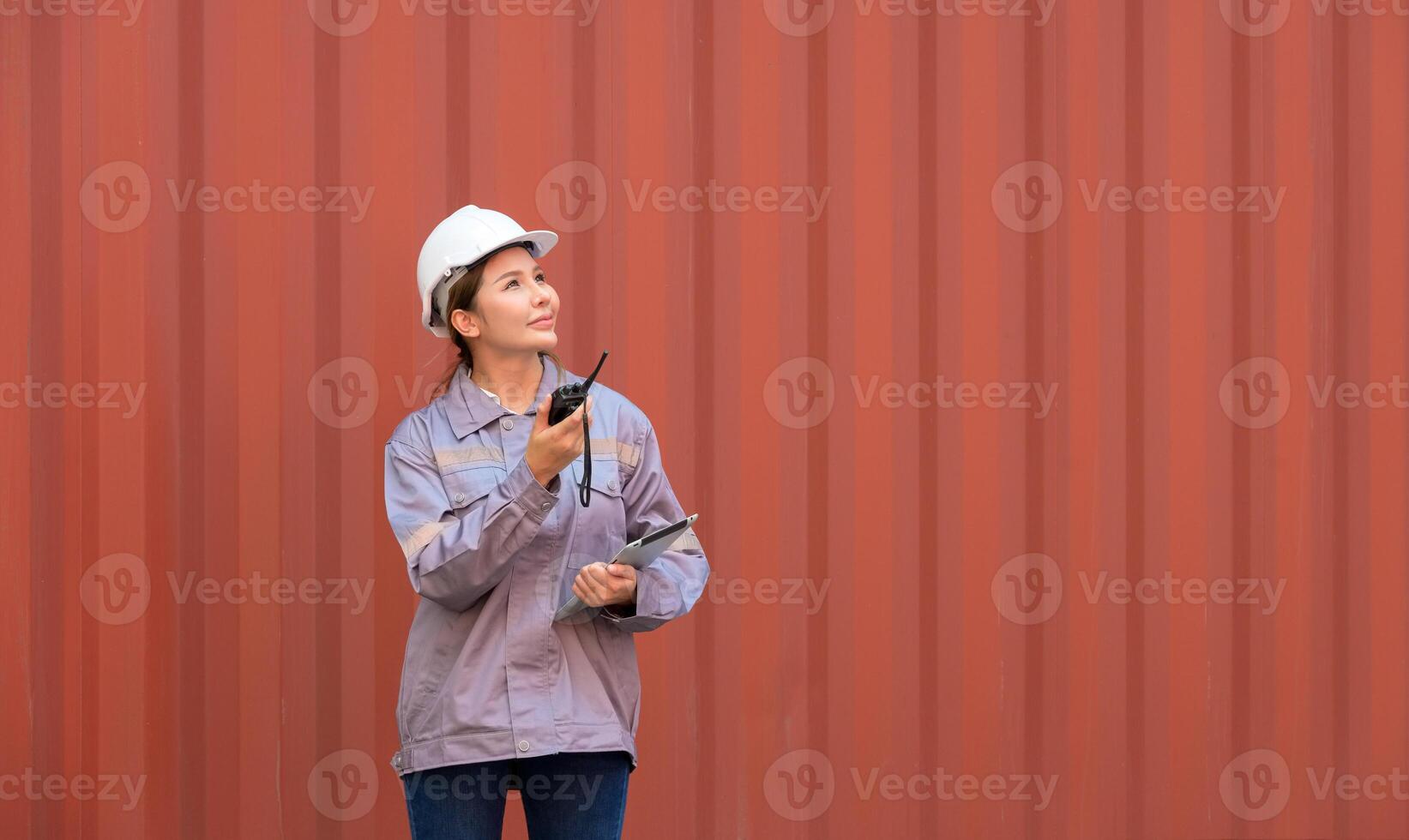 Female port worker in white helmet and uniform holding walkie talkie and tablet for coordinating logistics loading operations at container terminal shipping yard photo