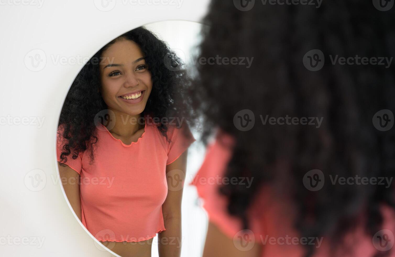 Beautiful dark skinned smiling girl in orange t-shirt looking at mirror admiring herself at home photo