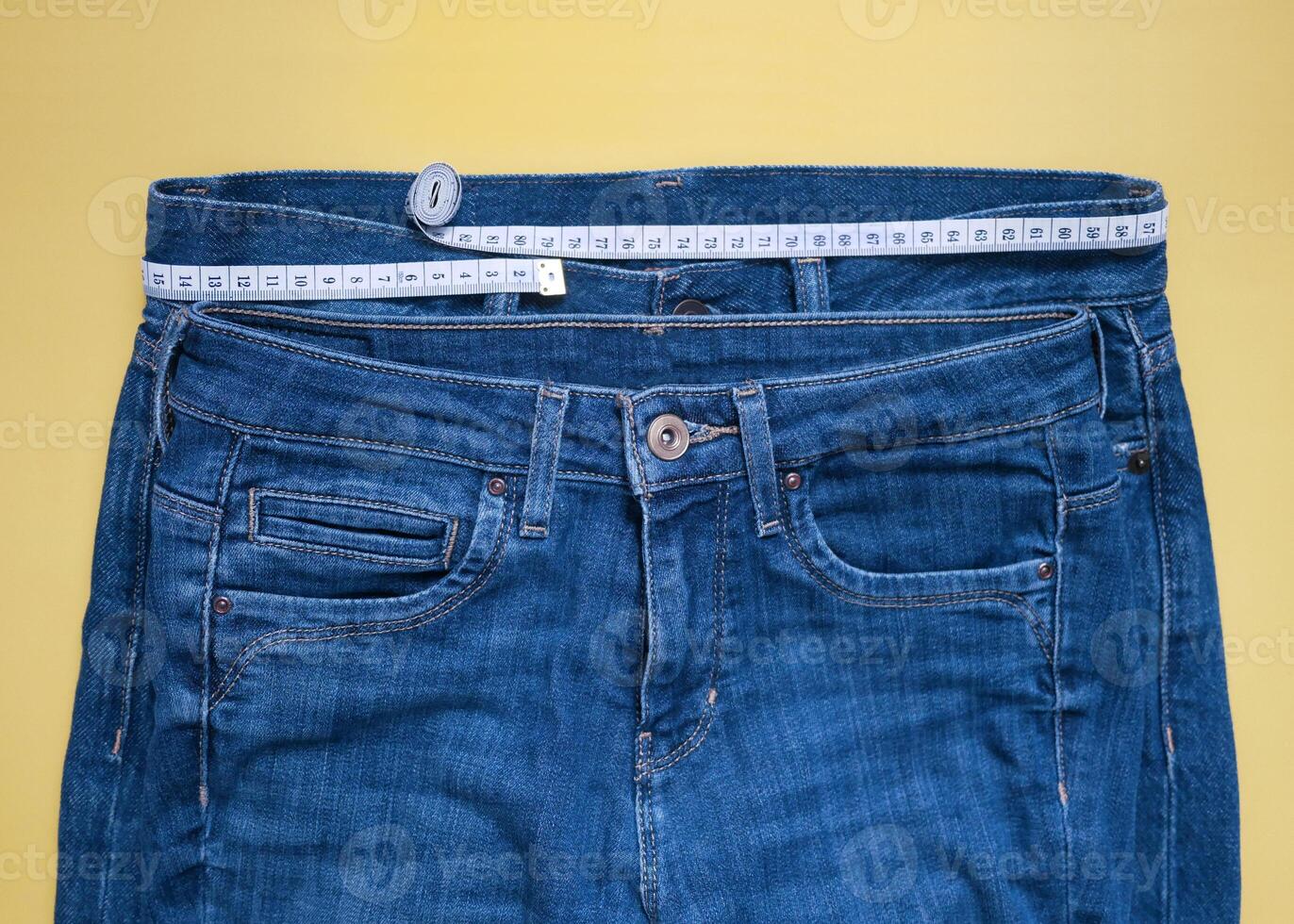 Two blue jeans of different size with measuring tape flat lay on a yellow background. Losing weight, overweight and healthy lifestyle concept photo