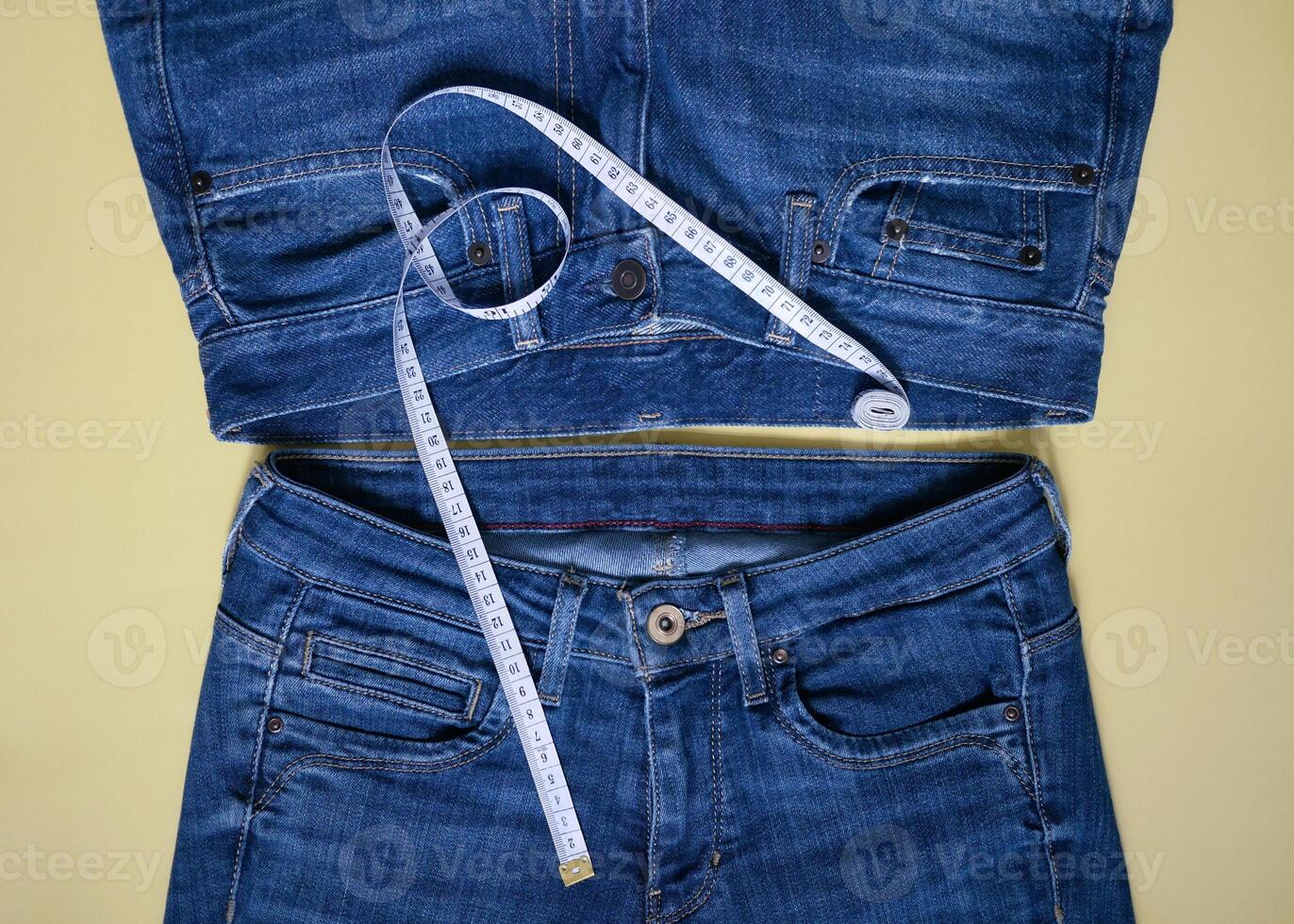 Two female blue jeans of different size with measuring tape flat lay on a yellow background. Losing weight, overweight and healthy lifestyle concept photo