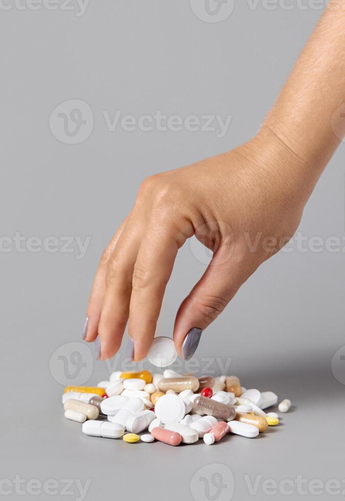 Woman hand put a pill on a pile of medical pills, tablets and capsules closeup against grey photo