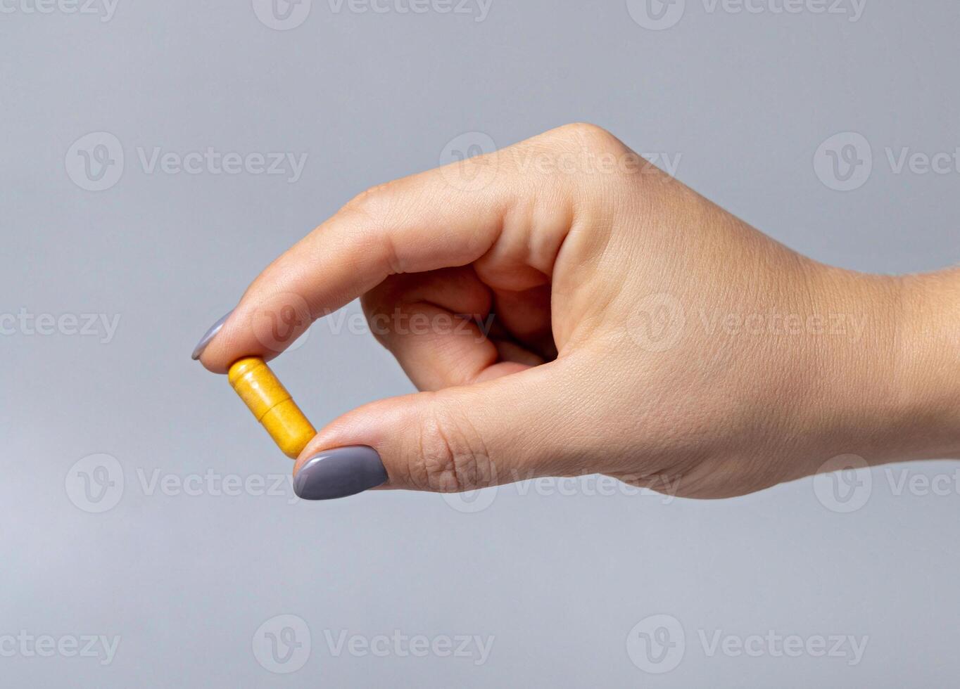 Female hand holding a yellow medical capsule with fingers closeup against grey photo