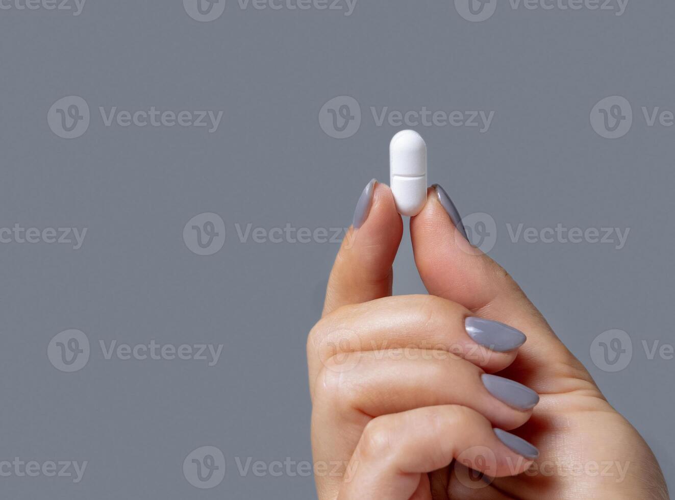 Female hand holding a white pill with fingers closeup against grey, copy space, Healthcare photo