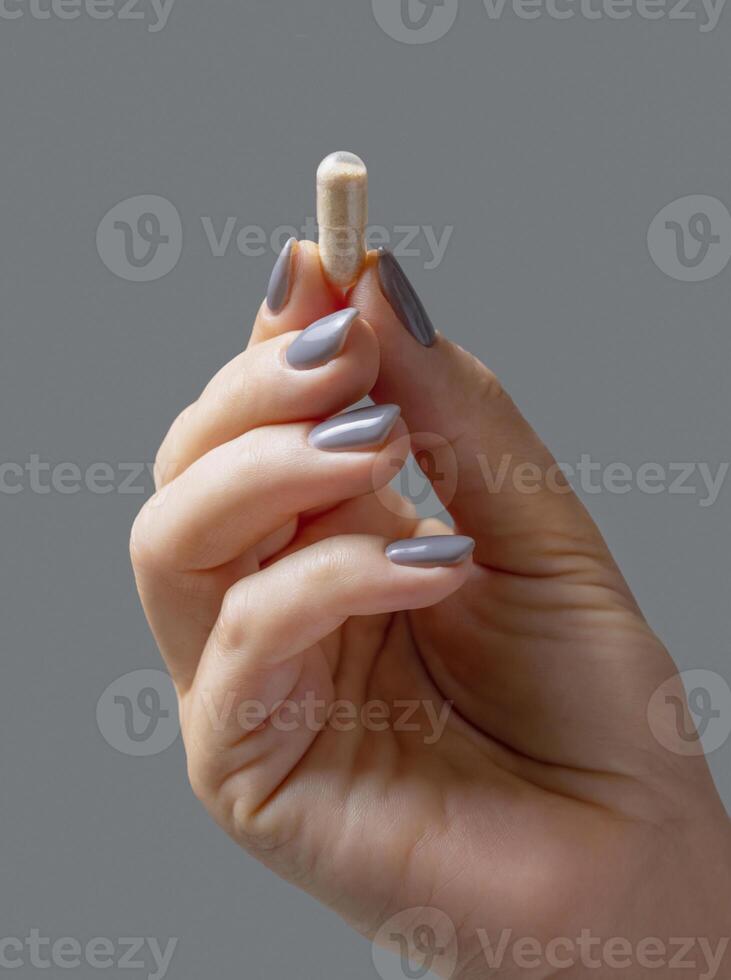 Female hand holding a beige capsule with fingers closeup against grey, Healthcare photo