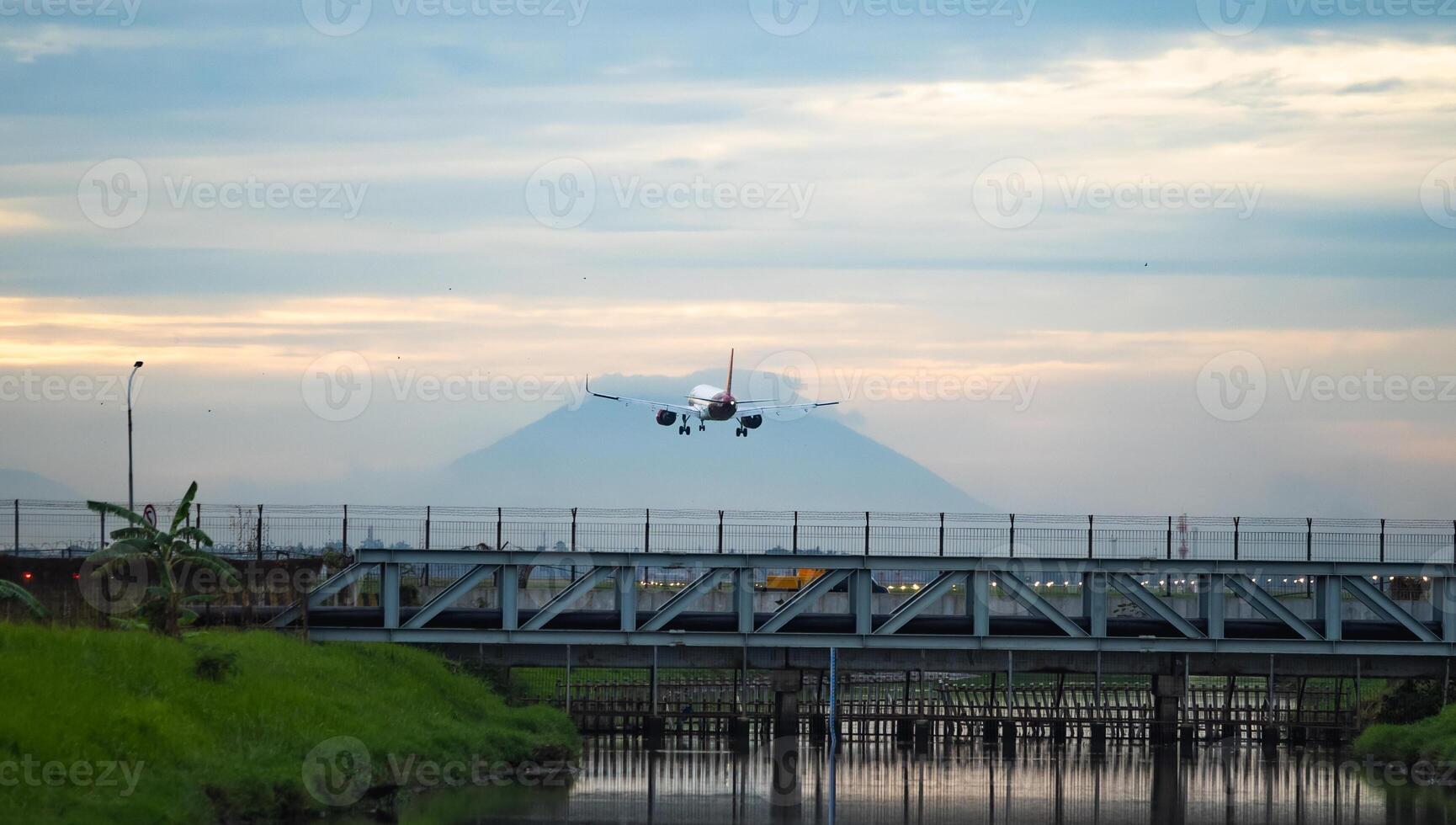 Airplane landing over a bridge and river at sunset with mountain silhouette photo