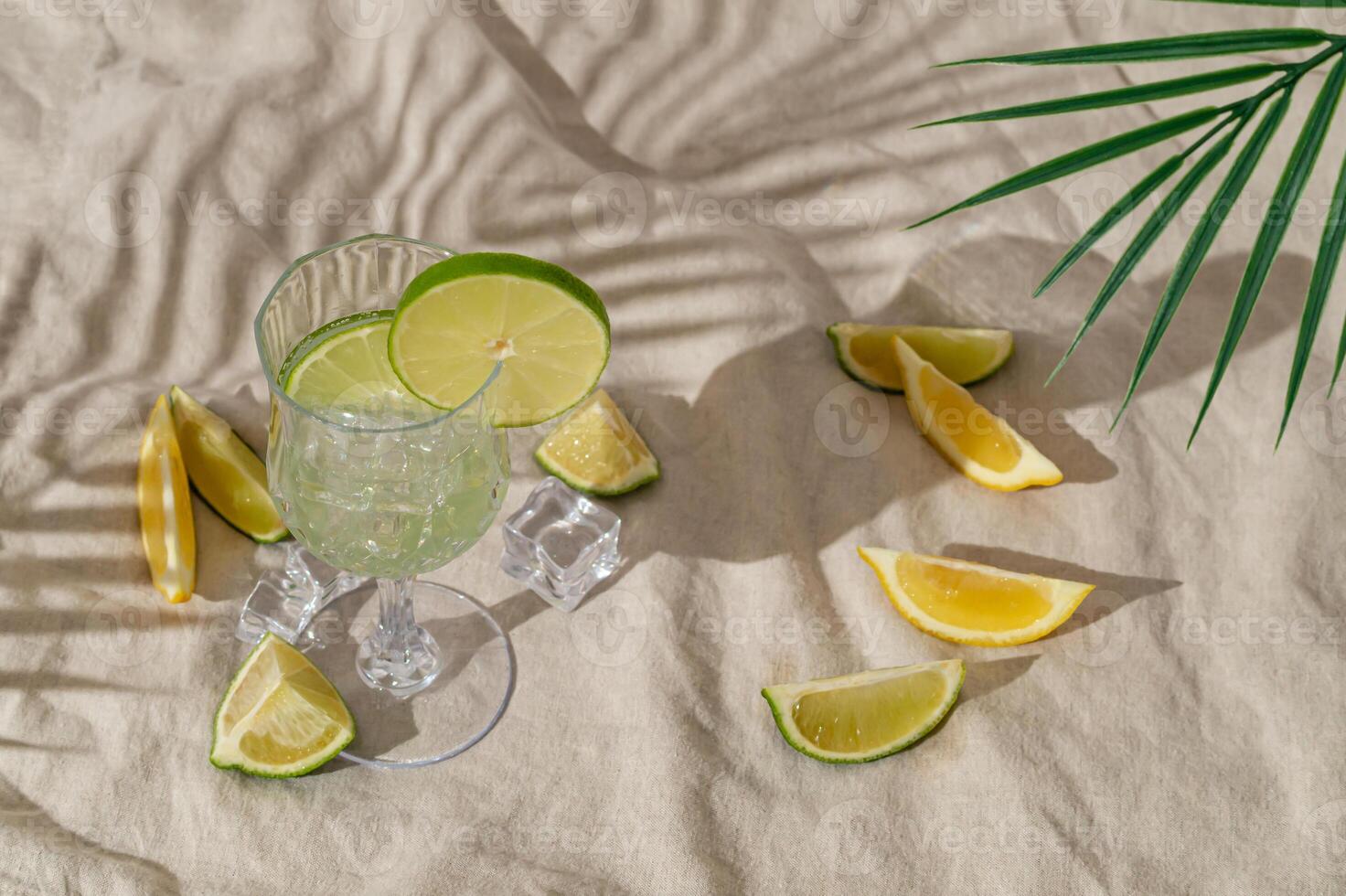 Summer day scene with refreshing drink, lime and lemon slices and tropical palm leaf shadow on sand color linen background. Sun and shadows. Minimal cocktail concept. Trendy summertime party idea. photo