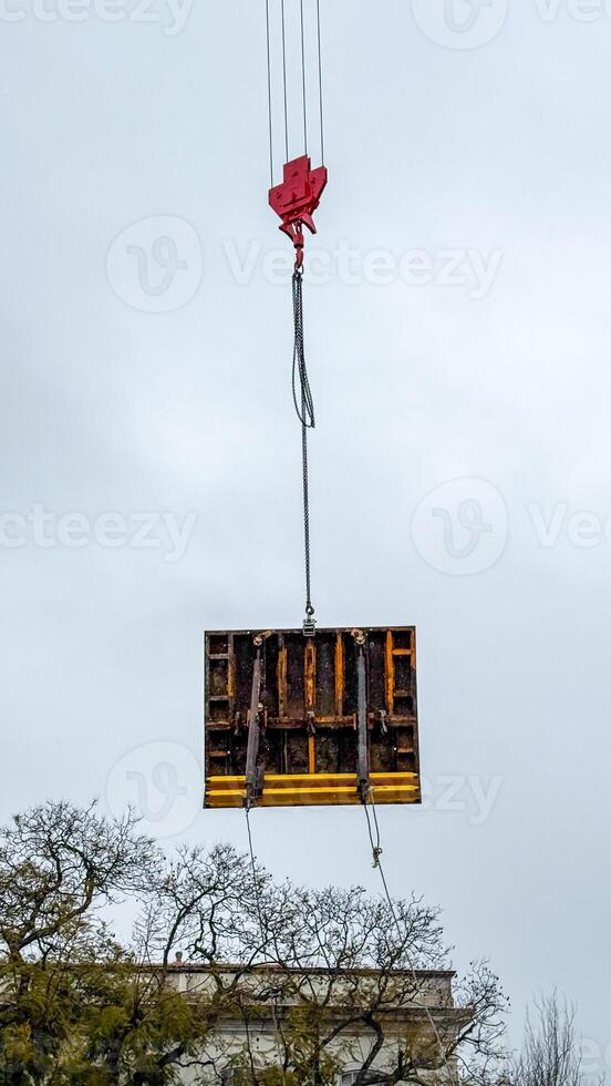Crane suspended pallet drifts like a modernist cloud above leafless branches, invoking International Workers Day industrial celebration photo