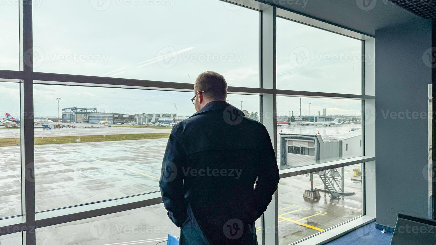 A contemplative Caucasian man gazes through rain kissed airport windows, embodying wanderlust and introspection, akin to World Tourism Day photo