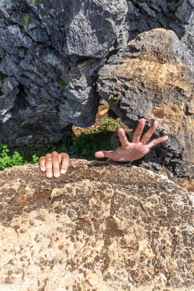 Two hands reach up from behind a rock formation, with an open palm, showing vulnerability and struggle. photo