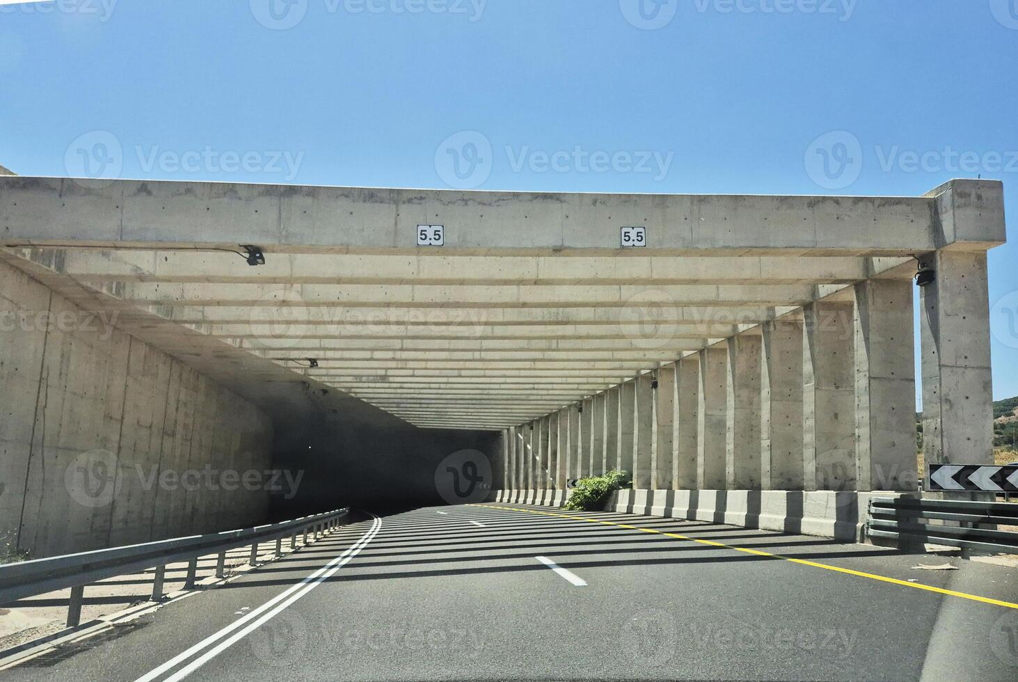 A modern concrete tunnel entrance with a 5.5m clearance height, casting strong shadows across the sunny mountain road photo