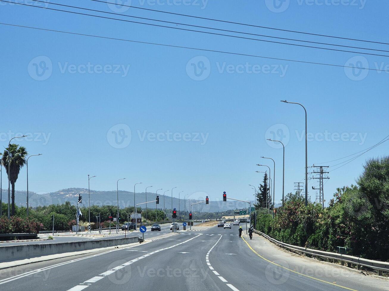 A wide open highway with traffic lights and clear skies, surrounded by greenery and distant hills under bright sunlight photo