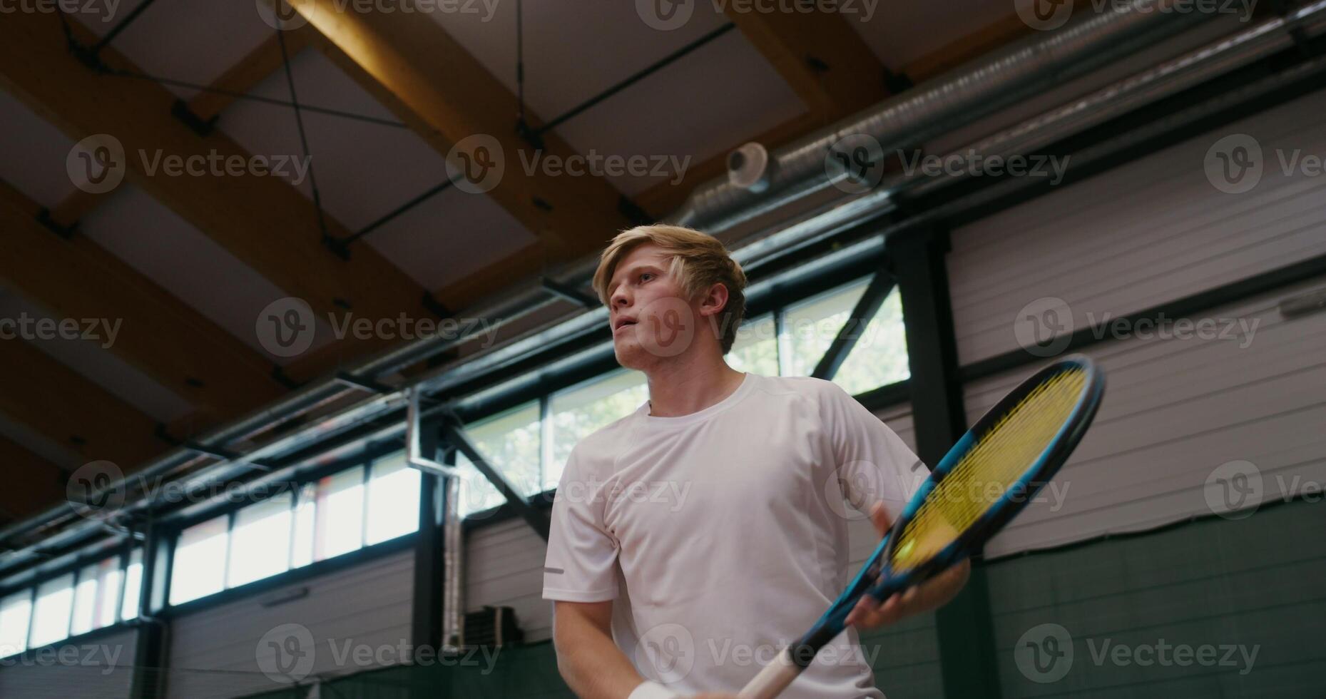 Young man of European appearance plays professional tennis, hitting ball after ball with a racket, playing on an indoor tennis court. Dynamic movements while playing tennis photo
