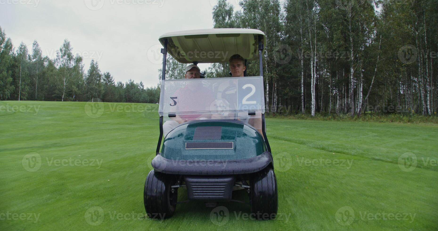 Men of European appearance with a big age difference are driving a golf car on a golf course and talking to each other on a rainy cloudy day, front view photo