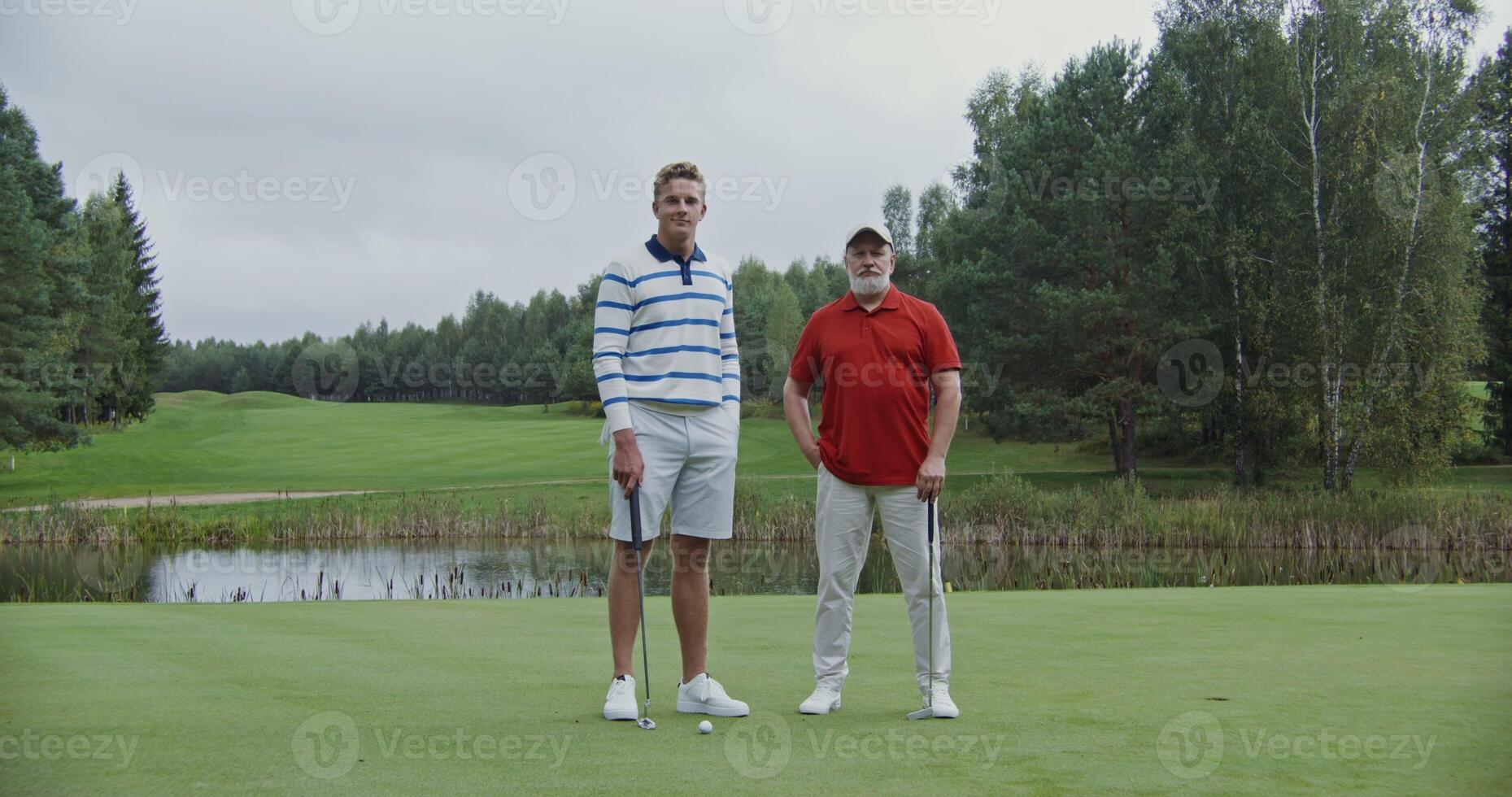 Two men of different ages are standing on the golf course with their legs crossed and smiling while looking at the camera. photo