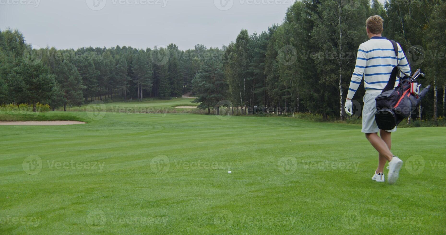 Young man in light-colored sportswear walks over to a golf ball, takes off his gear bag, grabs a golf club, and makes the next strike while watching the ball fly. photo