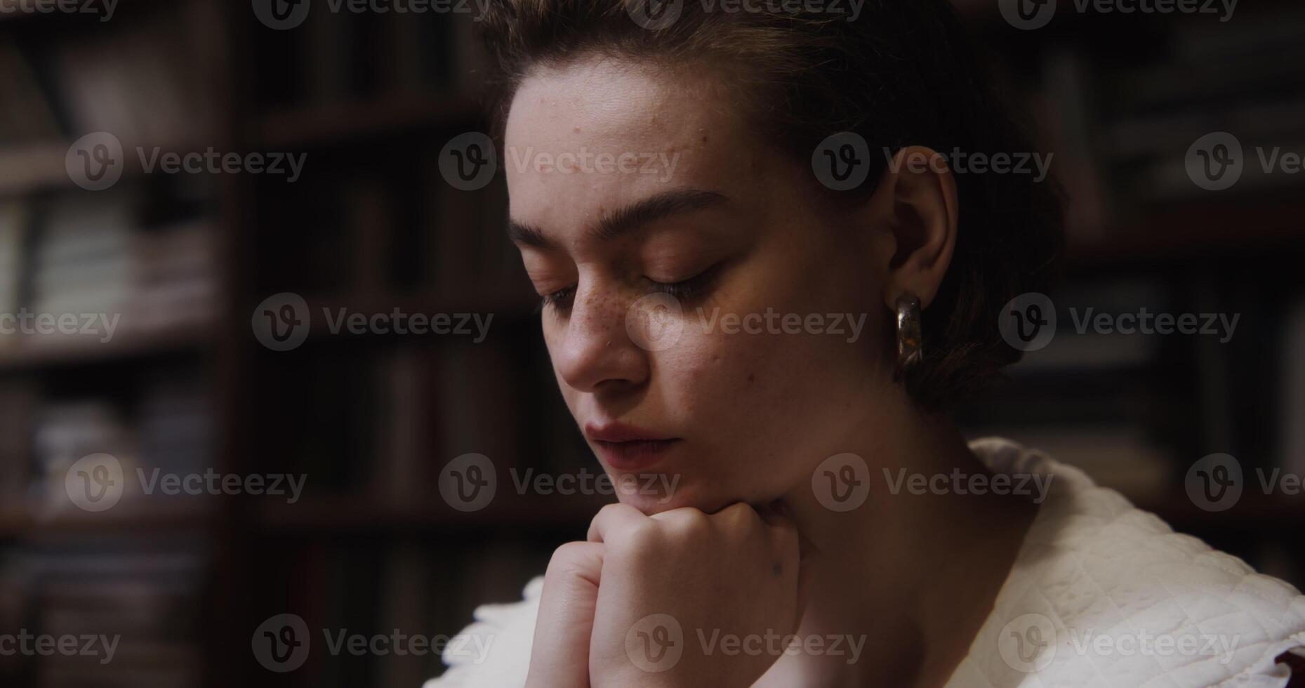 A beautiful girl of European appearance with freckles and in large earrings is thinking intently, resting her chin with her hands and looking down at the table, close-up photo