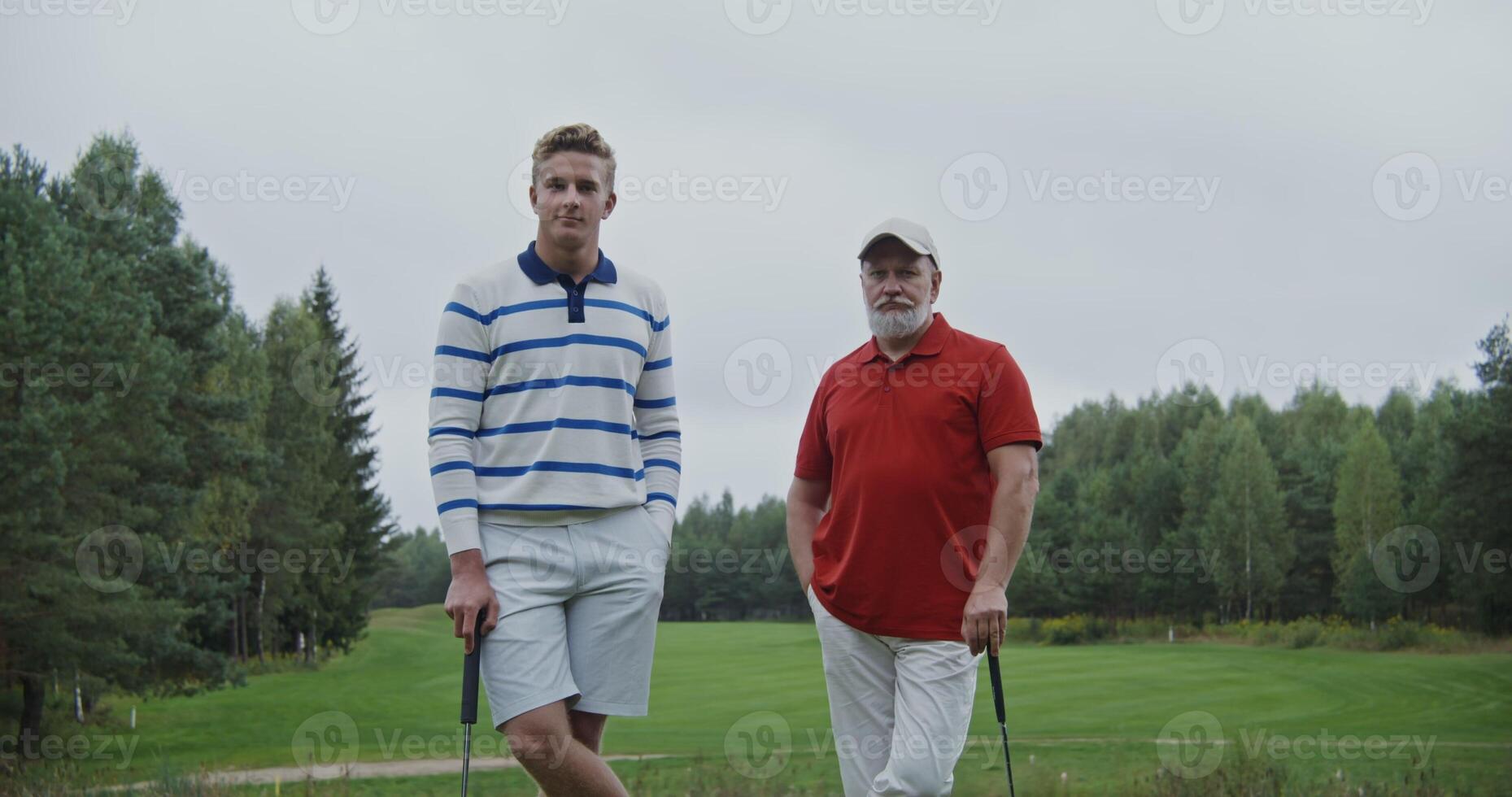 Two men of different ages are leaning on golf clubs on the golf course and smiling while looking at the camera photo