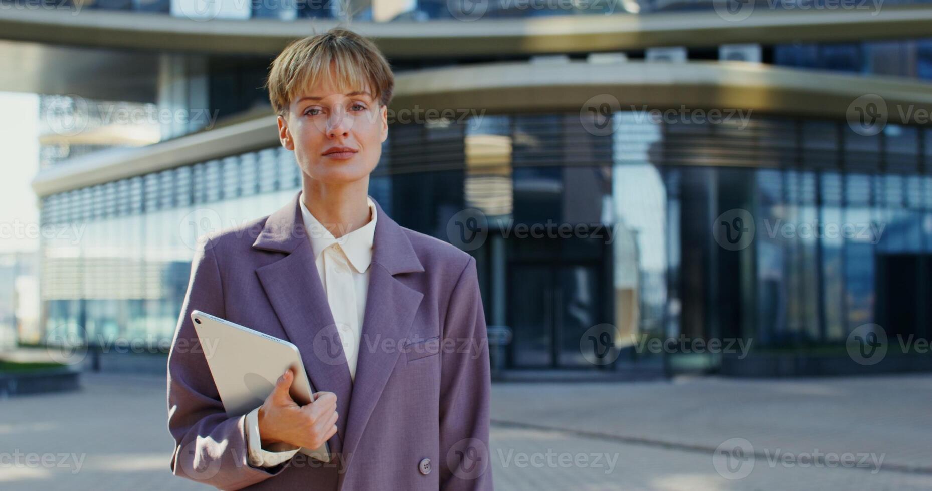 A beautiful young European woman with short hair, dressed in a business suit, holding a tablet and smiling while looking at the camera while standing outdoors near a modern office building. close up photo