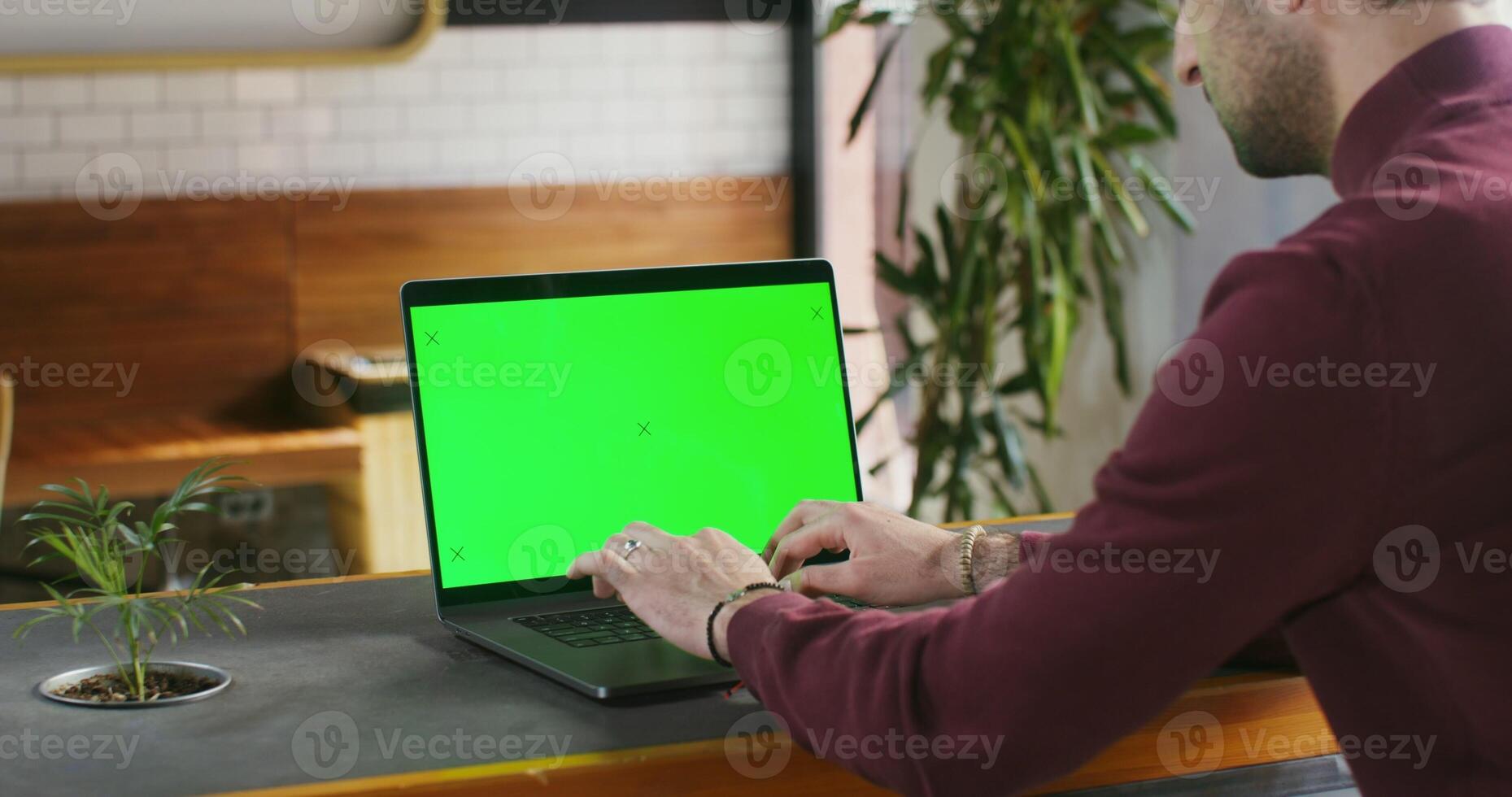 A young dark haired man with stubble on his face is typing on a laptop with a green screen while sitting at a table in a coffee shop. Chroma key, copy space. Videography from the side photo
