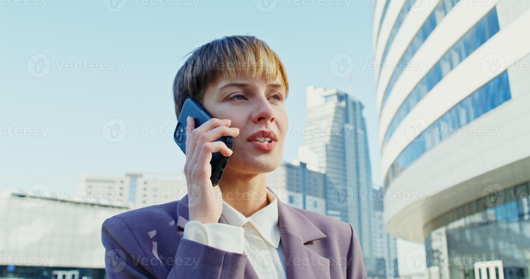 Beautiful young woman of European appearance with short hair, dressed in a business suit, smiling, talking on a mobile phone, standing near a modern office building, close up photo