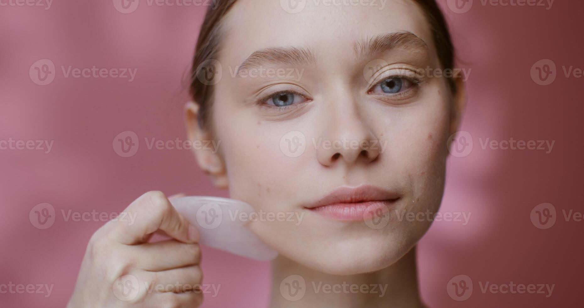 Beautiful young woman of European appearance makes a face massage with a pink jade stone Gua Sha and looks into the camera, standing on a solid pink background in the studio. 4K , red komodo photo