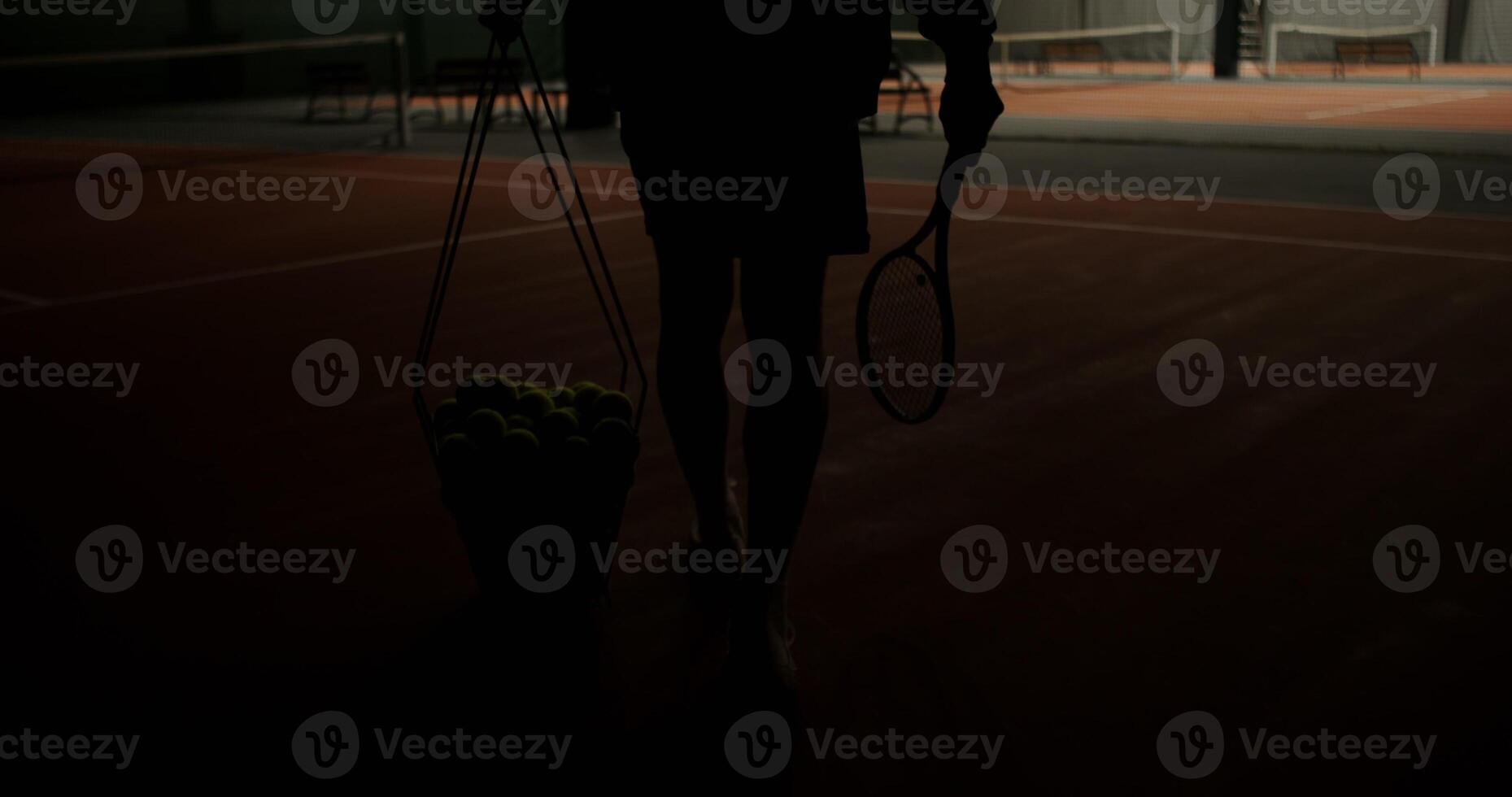 The tennis court lights come on and a man comes in, getting ready for the game, carrying a basket full of tennis balls. Videography of male legs with a basket from behind, close up photo