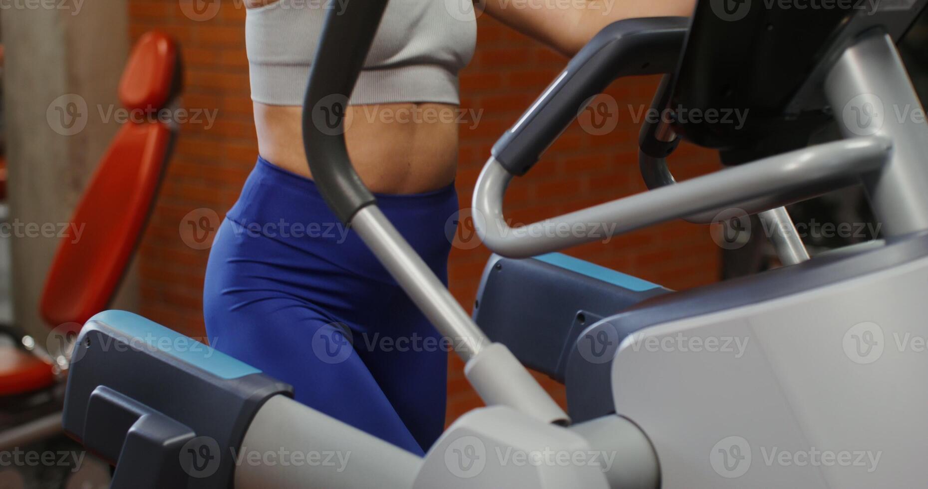 A young woman, dressed in sportswear, is engaged on a modern simulator for exercising in an indoor gym, a gradual displacement from the woman's legs to her face, close up photo