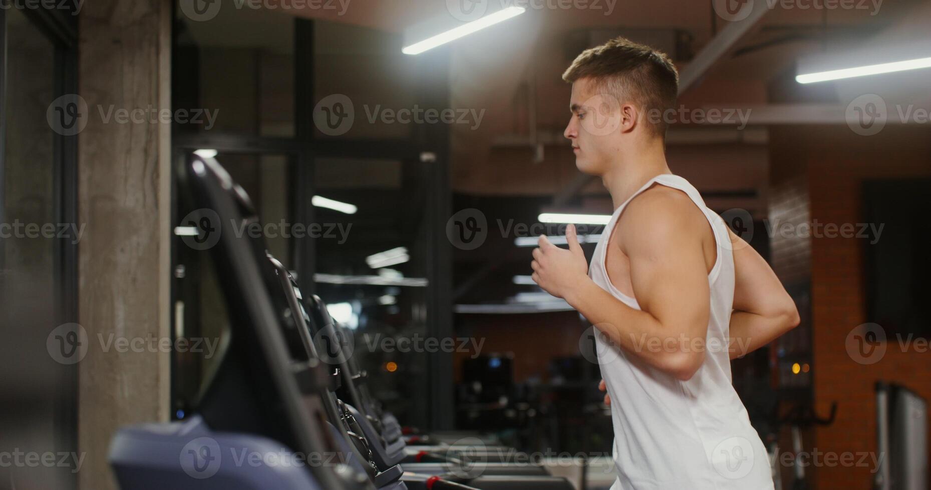 Young man of european appearance athletic physique trains on the running on a treadmill, exercising in a modern gym alone, close up, side view photo
