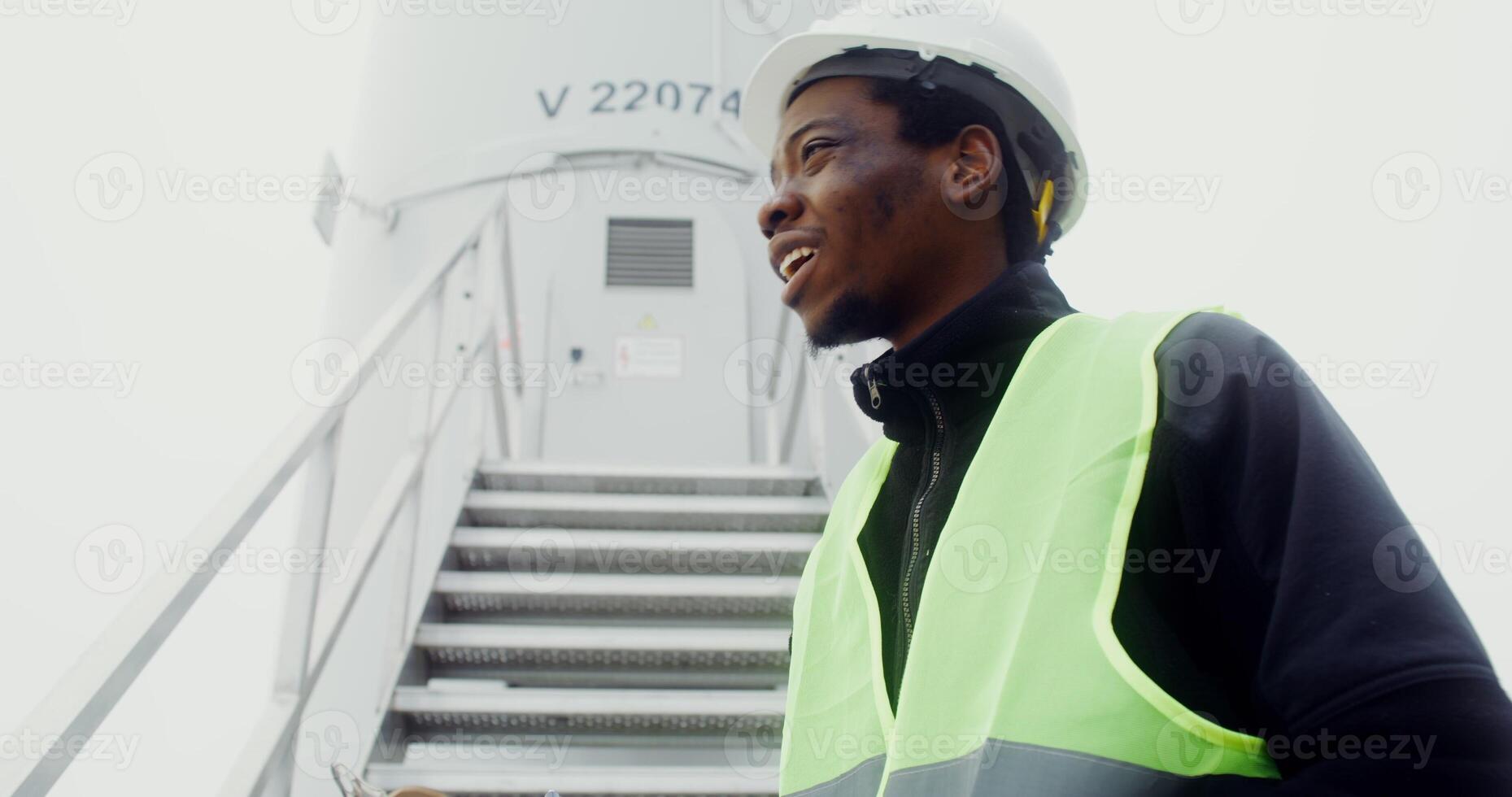 African American man in a green vest and hard hat takes notes in a glider while standing near the entrance to an industrial wind turbine, close up, dynamic . in 4k, red komodo photo