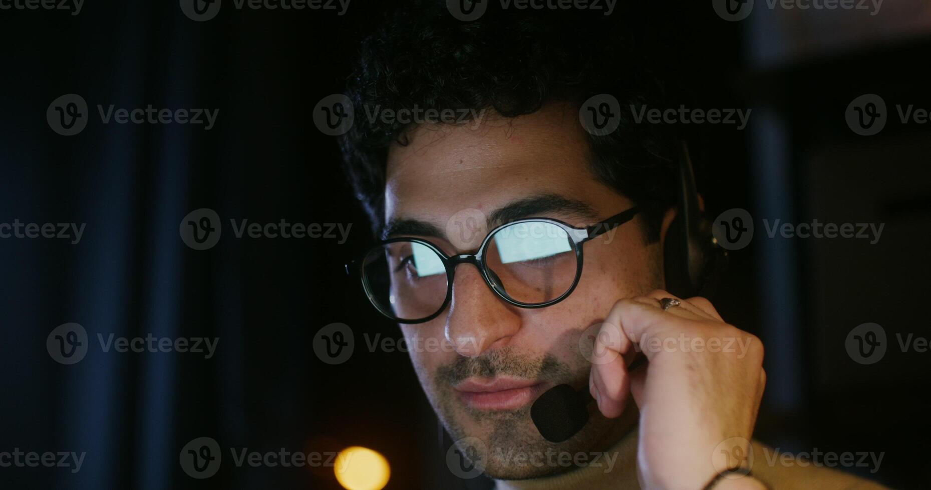 Young curly haired brunette man, wearing glasses, talking on a microphone with headphones and looking at a laptop in the dark, close up. Customer support photo