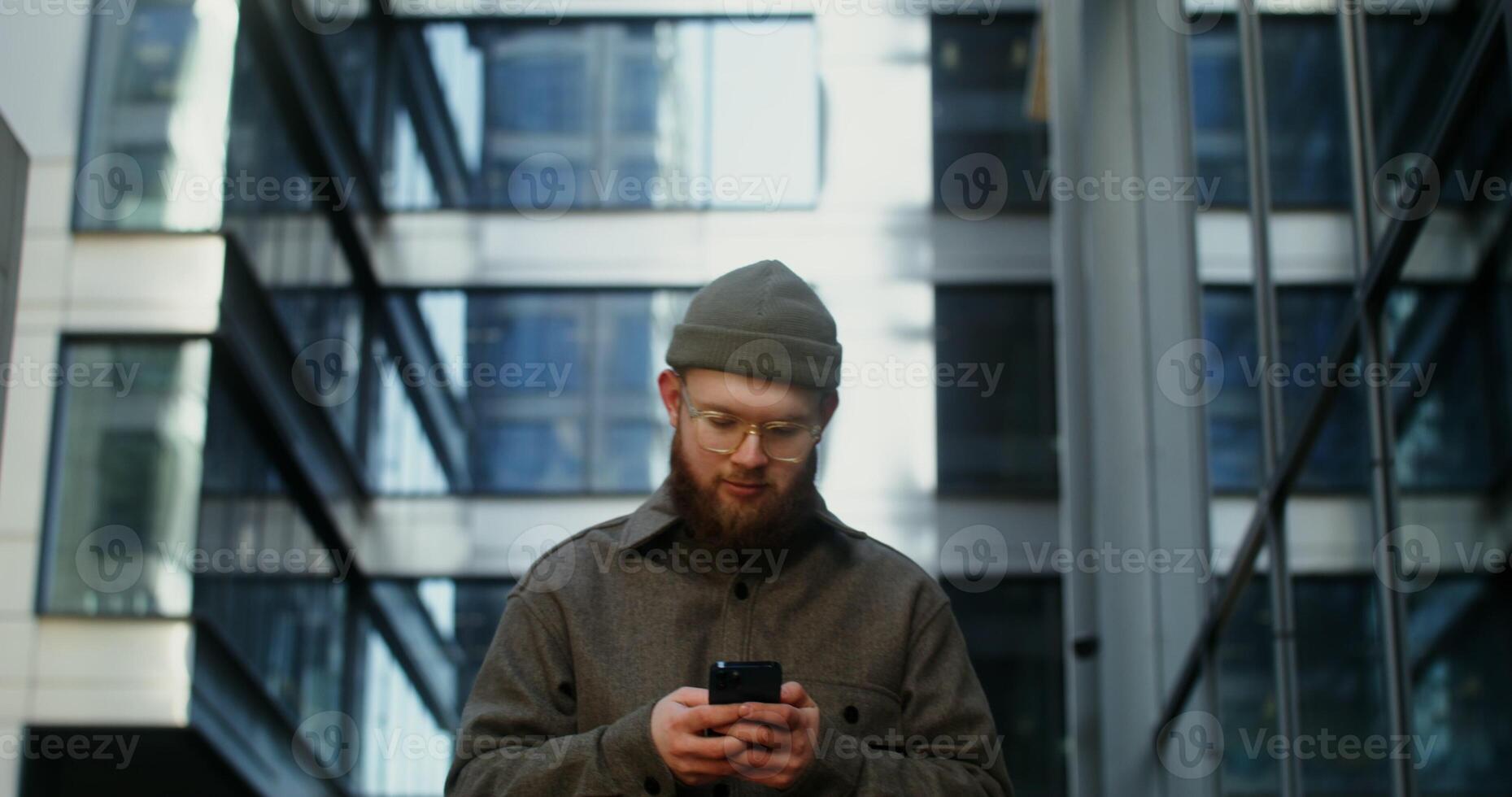 A young stylishly dressed man in a knitted hat is typing text on a mobile phone, walking around the business center of the city among office buildings, close up. in 4k, red komodo photo