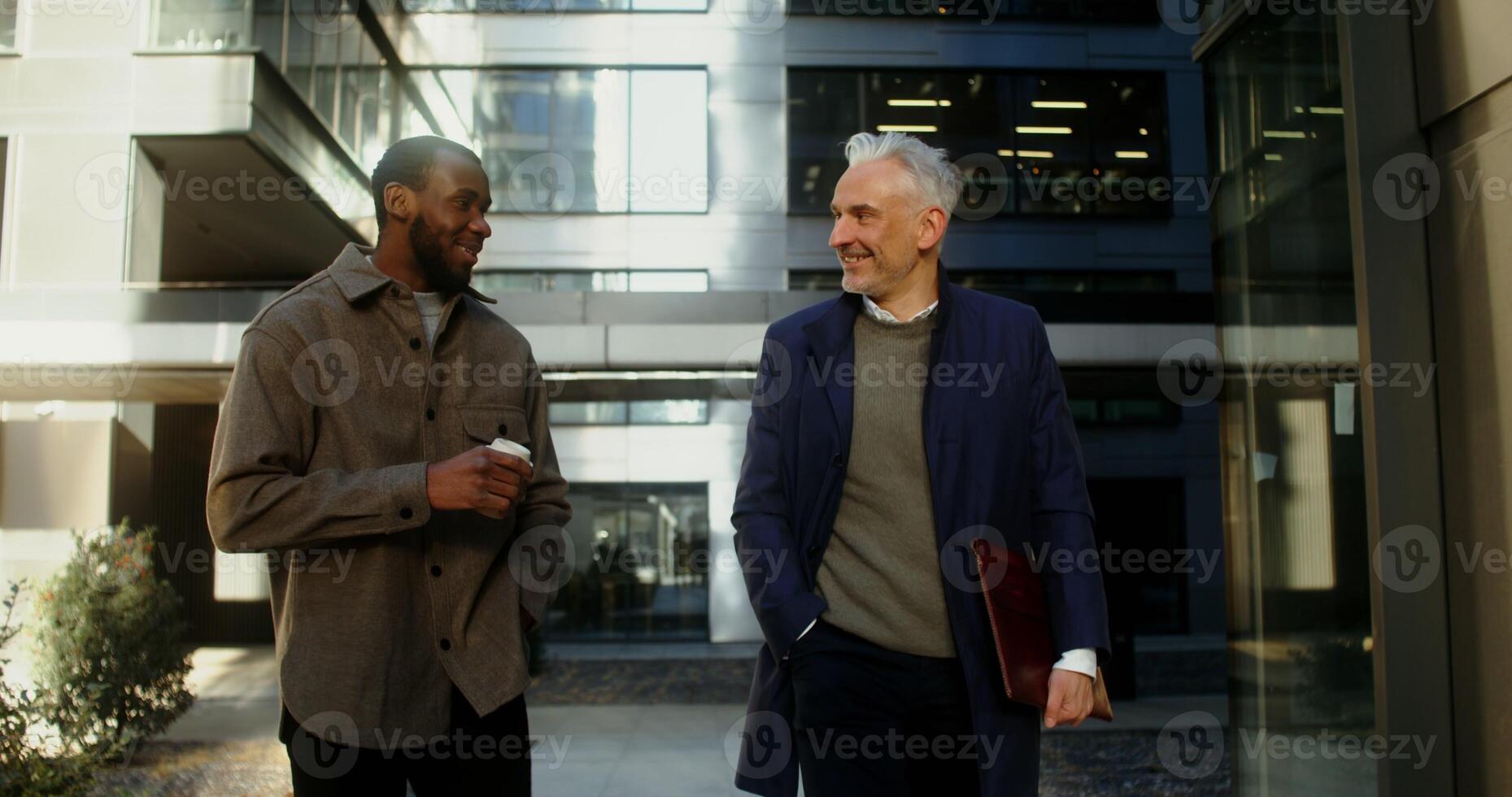 An African American man with a cup of coffee and a gray haired Caucasian man with a laptop are smiling while talking in the business center of the city on a sunny day. in 4k, red komodo photo