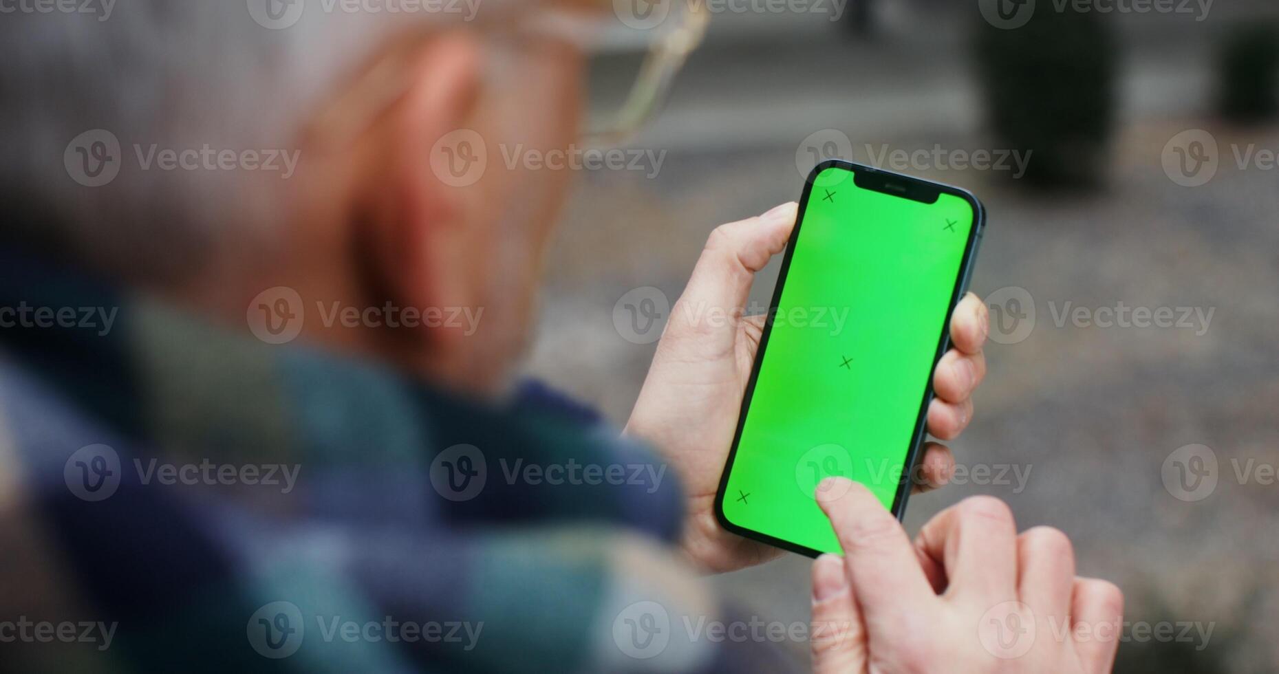 A gray haired man in a voluminous scarf uses a mobile phone with a green screen, an unrecognizable person, a top view from behind the man's shoulder. Chroma key, mockup. in 4k, red komodo photo