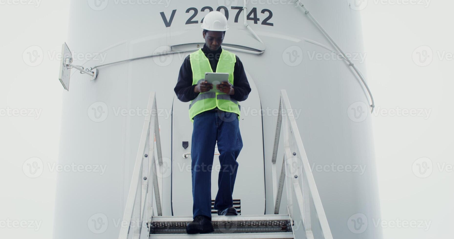A young African American man in work gear uses a tablet with a serious look as he walks down the steps of a wind turbine during an inspection. in 4k, red komodo photo