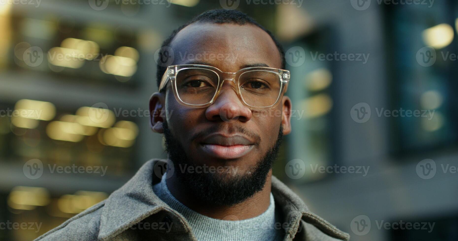 Stylish young African American man with a serious look at the camera, standing near a modern building in the evening, close up. Blurred windows with a lit light on background. in 4k, red komodo photo