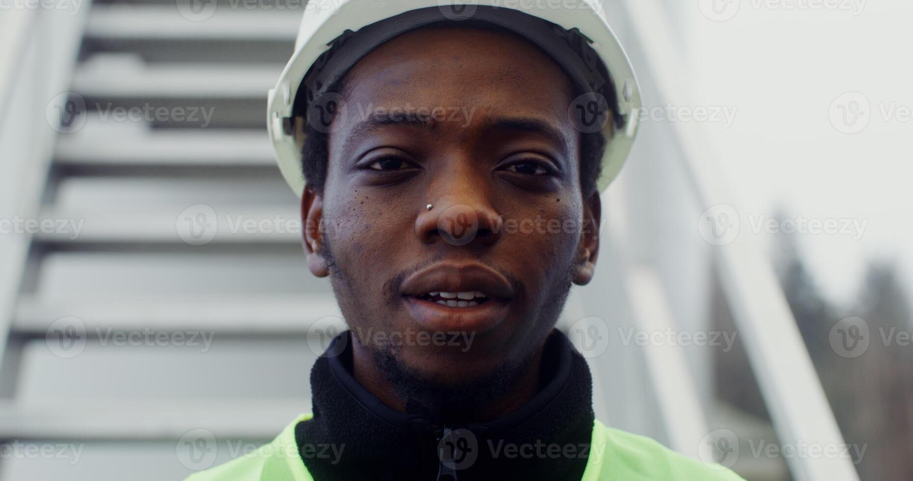 A young African American man in a white hard hat and a green vest looks at the camera with a smile, close up. in 4k, red komodo photo