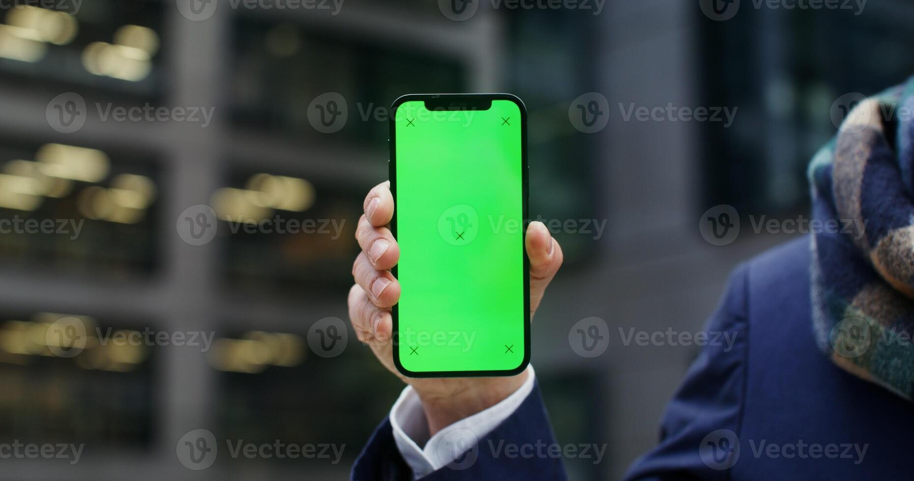 A warmly dressed man holds a green screen phone as he stands outside an office building. Copy space, mockup, close up of the phone, no face. in 4k, red komodo photo
