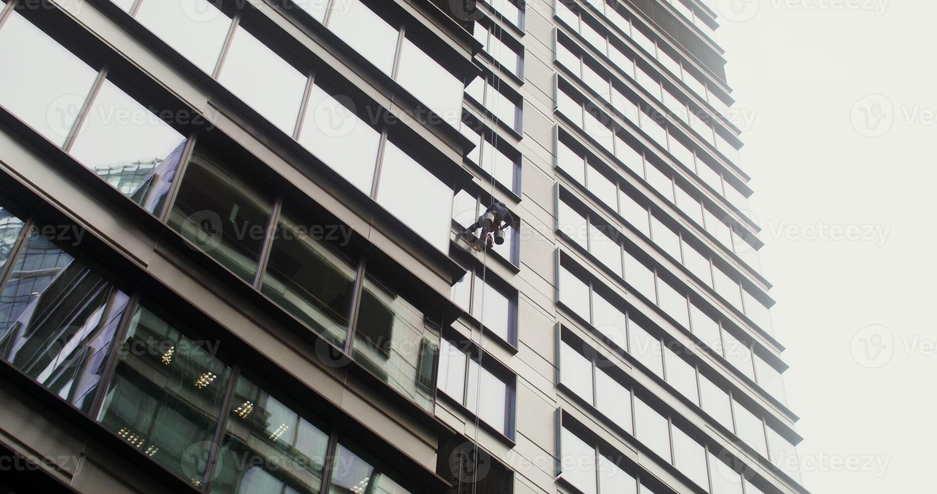 A man in a protective helmet and gear hanging from protective cables washes the windows from the outside of a high rise building. Industrial cleaning. in 4k, red komodo photo