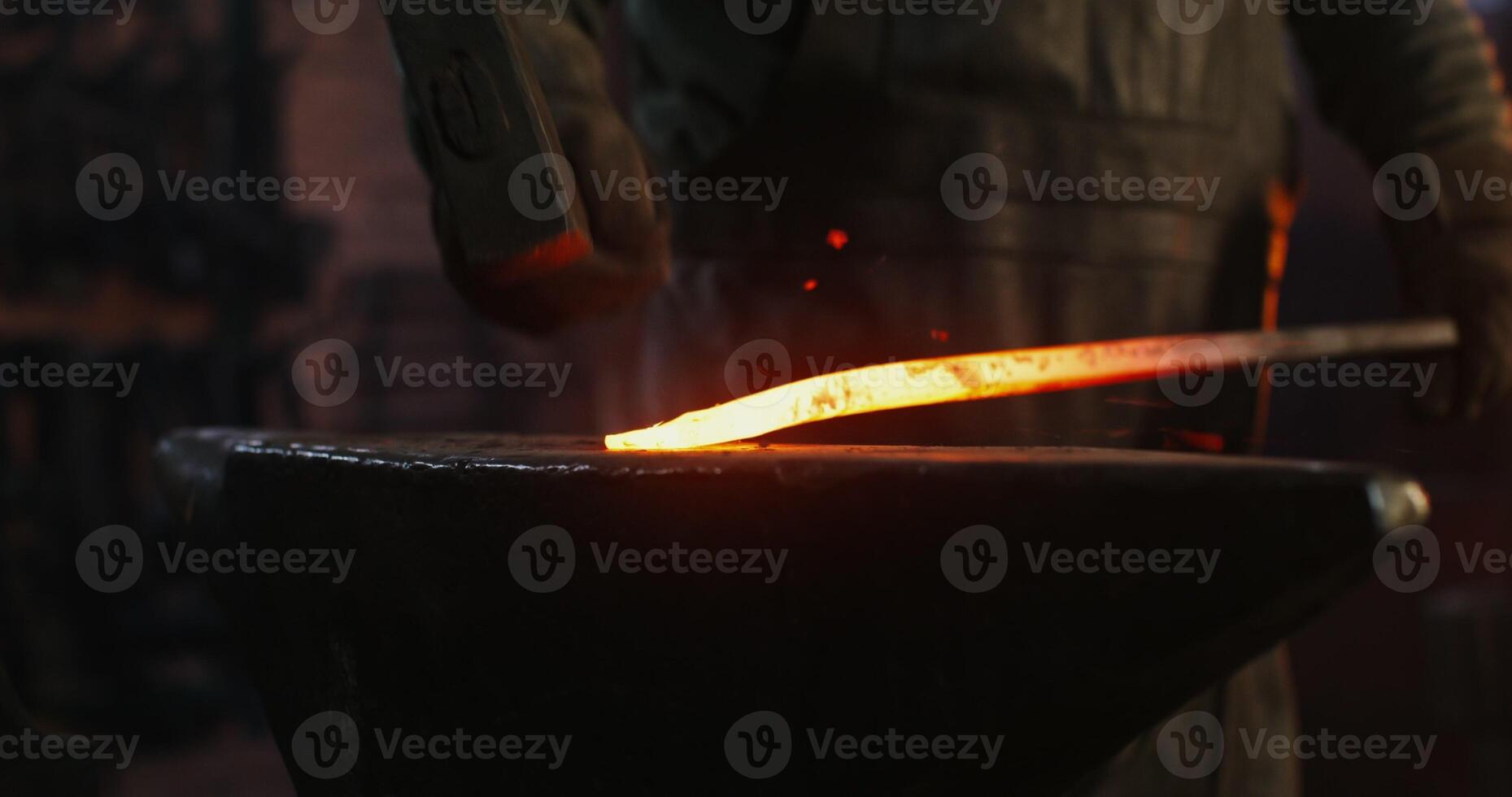 A blacksmith in protective gear takes out a red hot metal billet from the furnace and begins to shape it with a hammer, close up, no face. Work in a modern forge. 4k , red komodo photo
