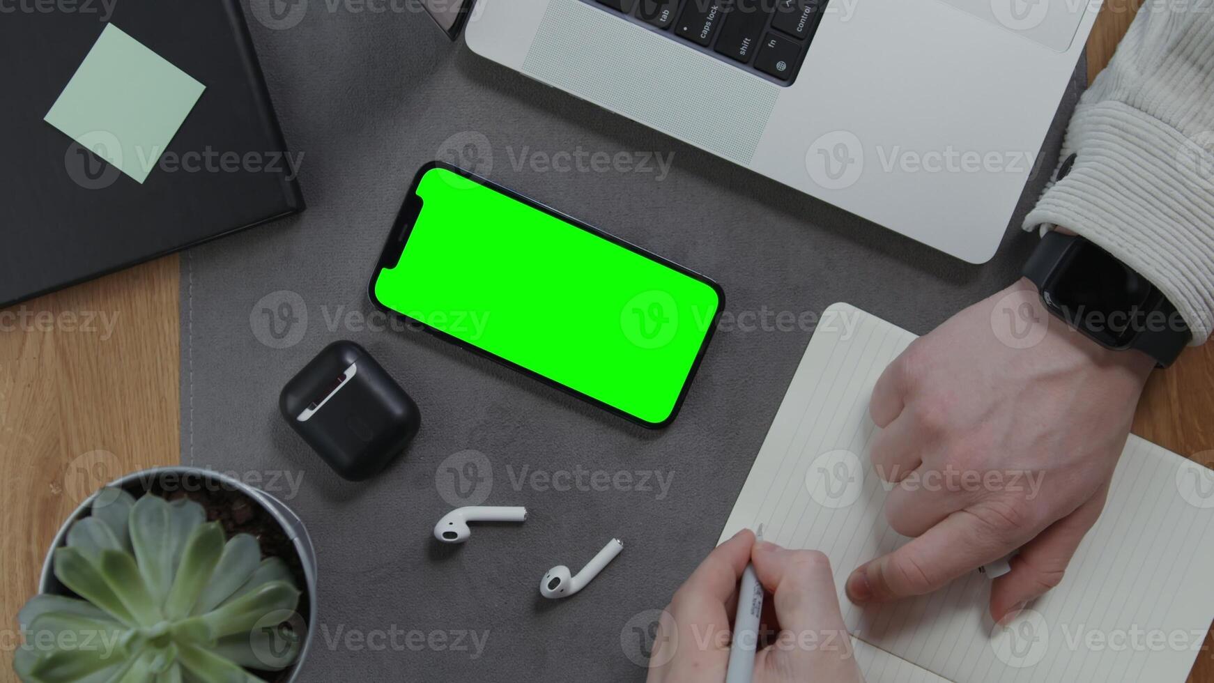 A man makes notes with his left hand in a notebook, lying next to a mobile phone with a green screen on the desktop, close up of male hands. Mockup, chroma key, copy space photo