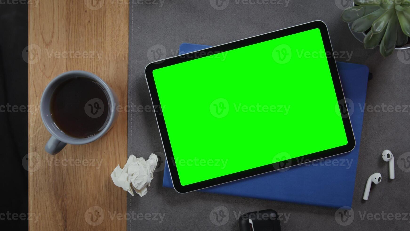A digital tablet with a green screen lies on the desktop next to a cup of hot tea, top view, close up, no people. Mockup, chroma key, copy space photo