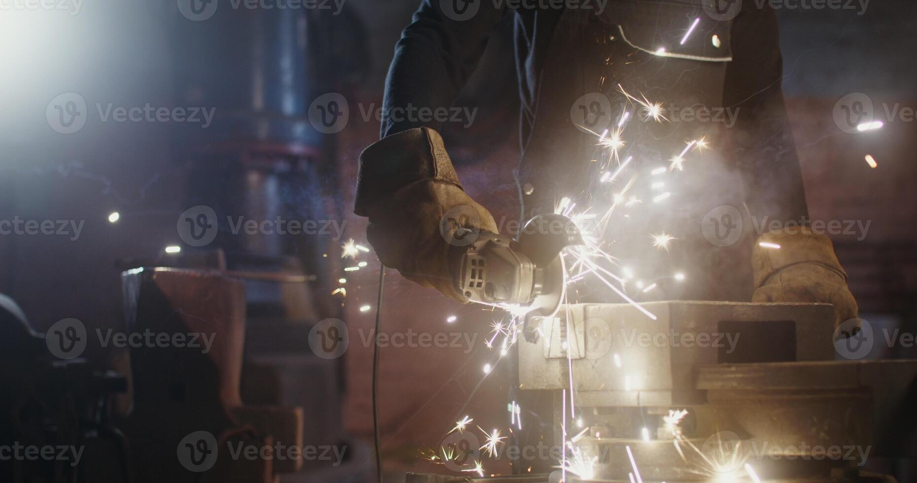 A blacksmith of African American appearance in goggles and an apron cuts off a piece of metal workpiece using an angle grinder. Close up, motion from the man's hands to his face. 4k, red komodo photo