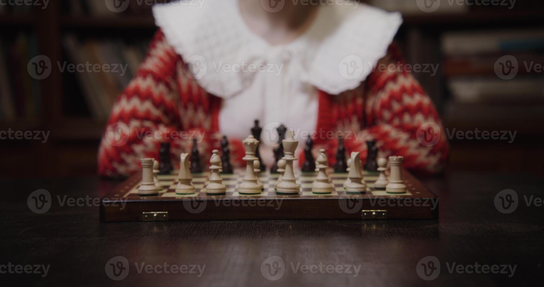A young woman of European appearance plays a chess game alone, thinking long concentratedly between moves, sitting at a table in the library, close up photo