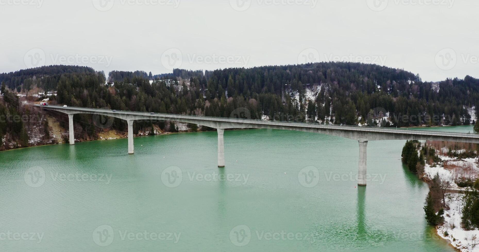 Minimalist concrete bridge for road transport across wide river in a picturesque place among forests and fields covered with snow, drone , from the side of the river photo