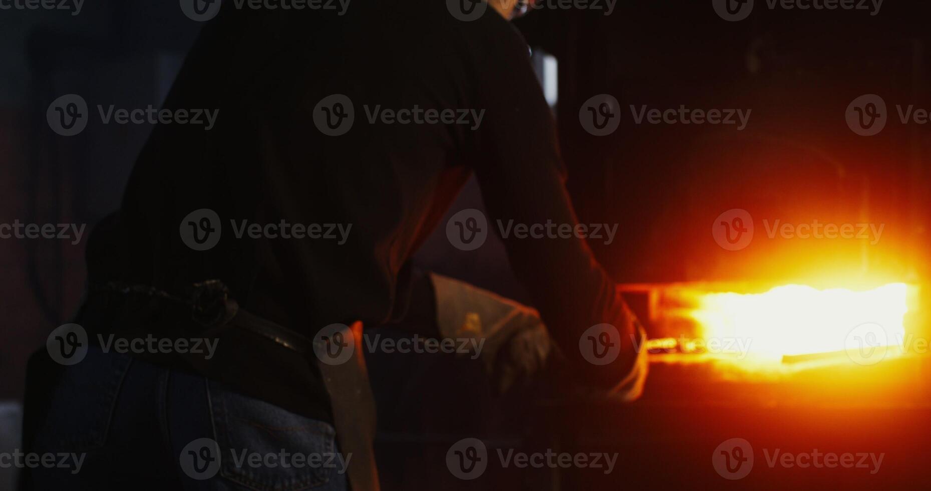 A young man in goggles and a leather apron takes a split metal billet out of the furnace with tongs and starts shaping the product with a hammer, close up. in 4k, red komodo photo