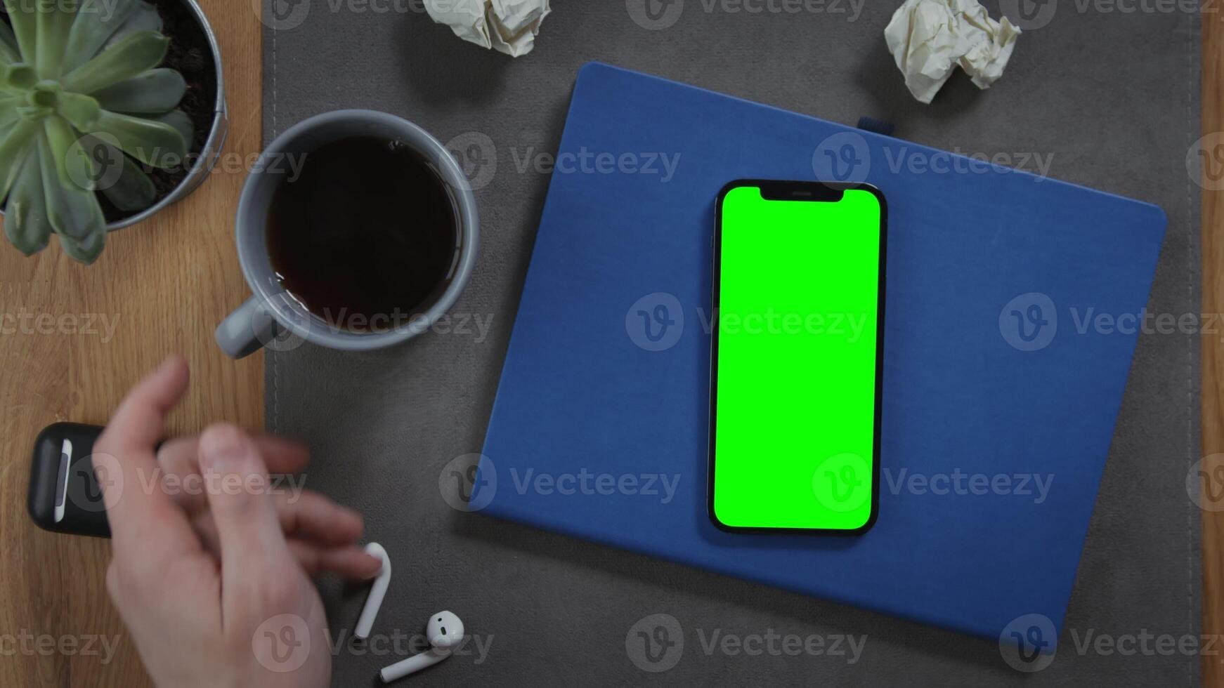 A man takes a cup of hot coffee, standing on desktop next to a mobile phone with a green screen, lying on a notebook with pieces of paper crumpled next to it, close up. Mockup, chroma key, copy space photo
