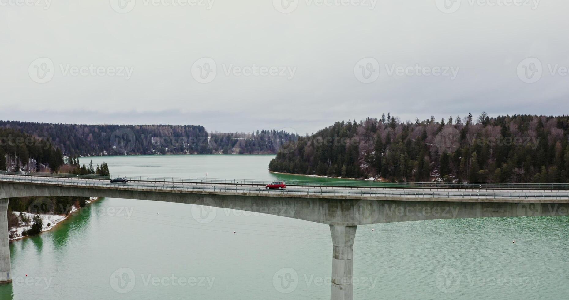 View of wide river flowing among spruce forests and fields covered with snow, and a narrow concrete road bridge connecting the banks, filming from a bird's eye view, from a drone photo