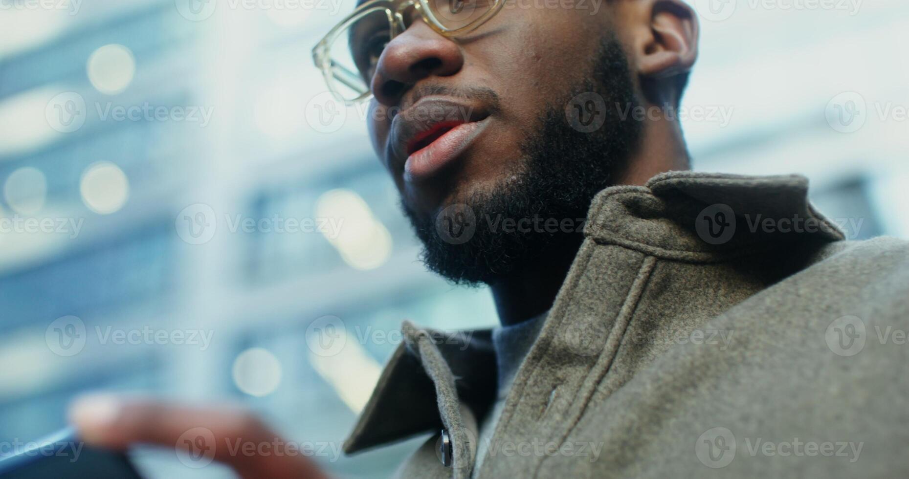 An African American man uses a mobile phone while standing near modern buildings. Close up of a focused male face, viewed from below. in 4k, red komodo photo